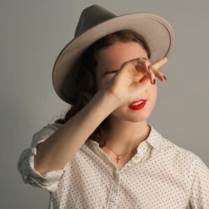 A woman wearing a grey fedora and a white polka-dot shirt, partially shielding her face with her hand, featuring bright red lipstick and a gold cross necklace.