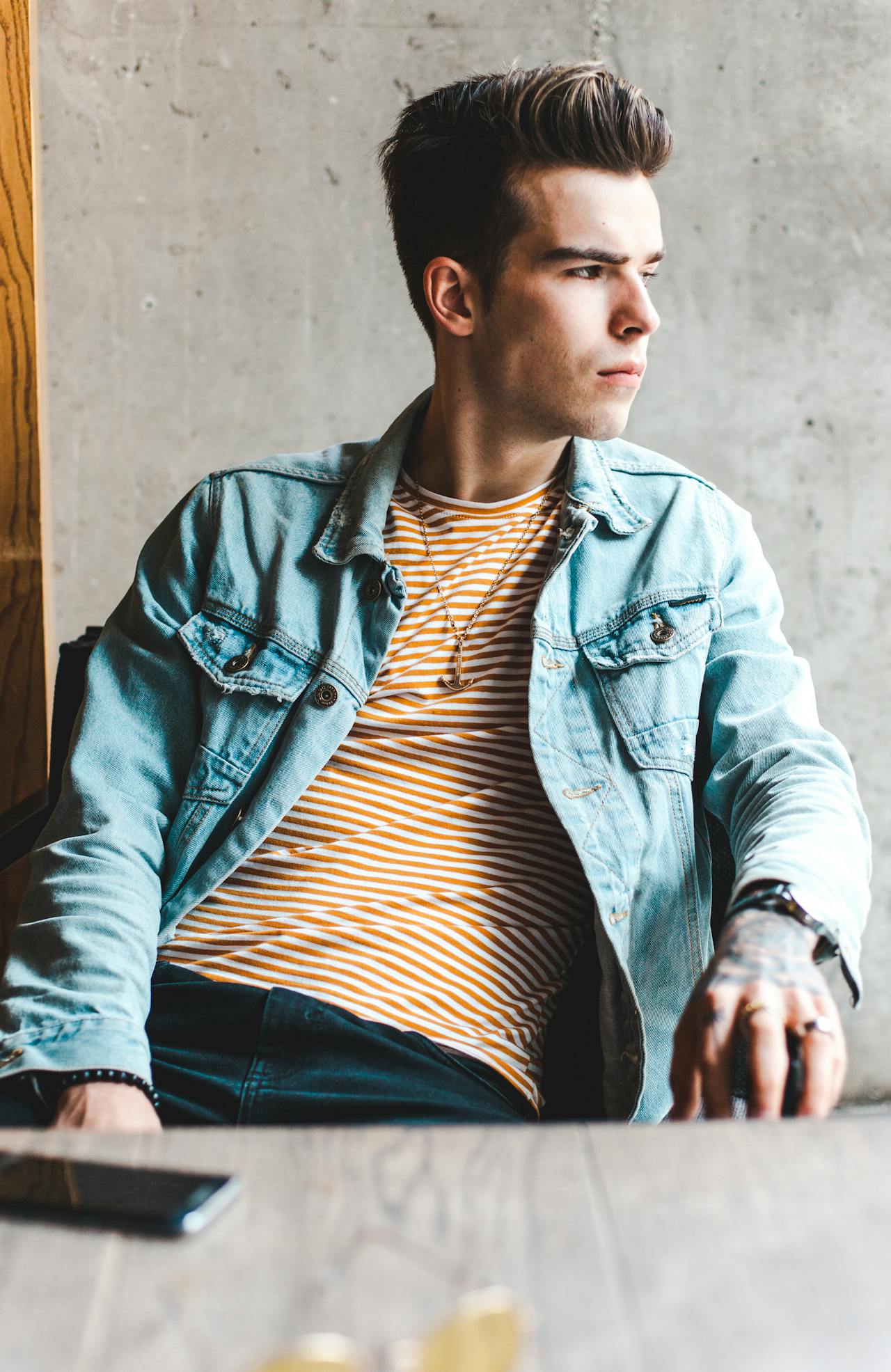 A young man with styled hair looking out a window, wearing a light blue denim jacket over a yellow and white striped shirt with an anchor necklace.