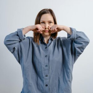 A woman with shoulder-length brown hair wearing an oversized blue button-down linen shirt, holding her fists to her cheeks in a playful or thoughtful pose against a white background.