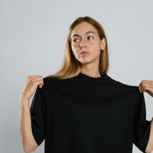 A young woman with long blonde hair holding the shoulders of a plain black oversized t-shirt, looking away against a light grey background.