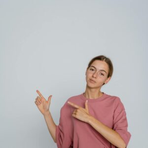 A young woman in a dusty rose oversized t-shirt and denim jeans pointing her fingers to the side against a plain white background.